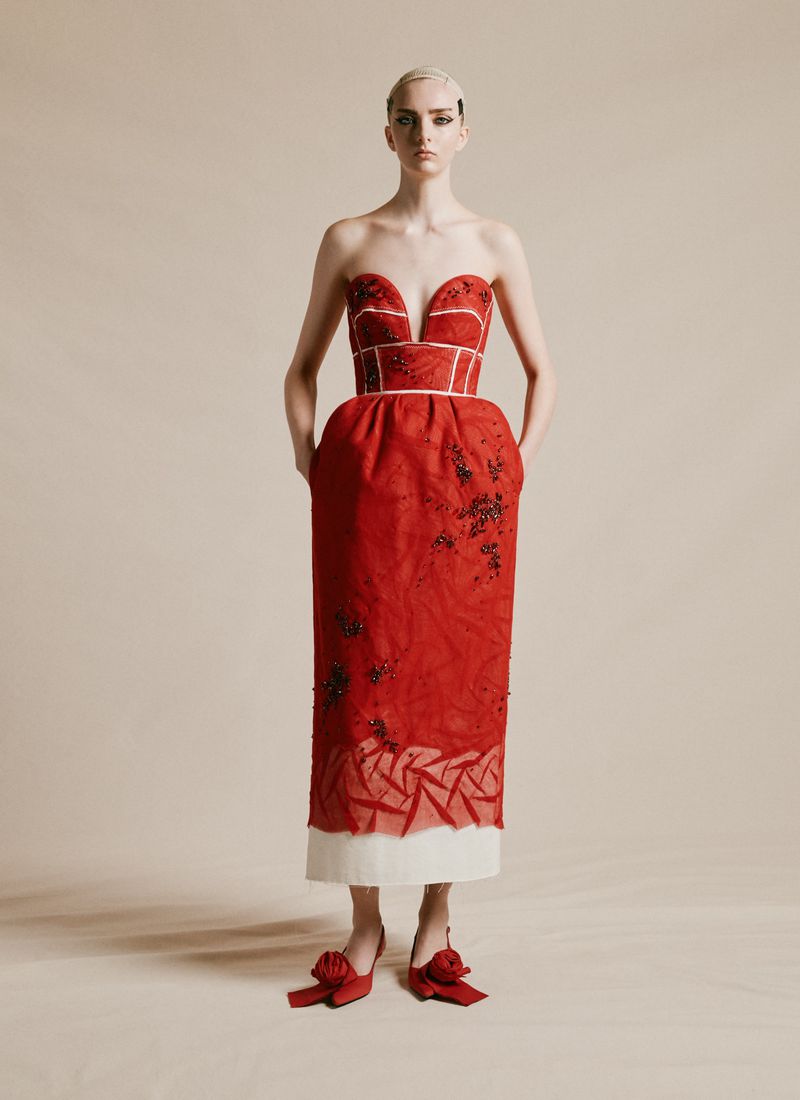 Full-length studio shot of a woman in a red strapless dress and red shoes, hands in pockets, against a neutral background.