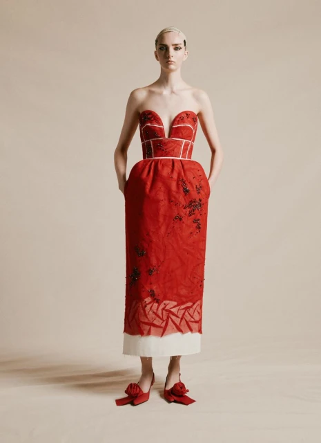 Full-length studio shot of a woman in a red strapless dress and red shoes, hands in pockets, against a neutral background.