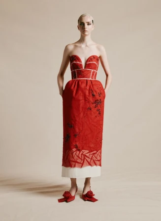 Full-length studio shot of a woman in a red strapless dress and red shoes, hands in pockets, against a neutral background.
