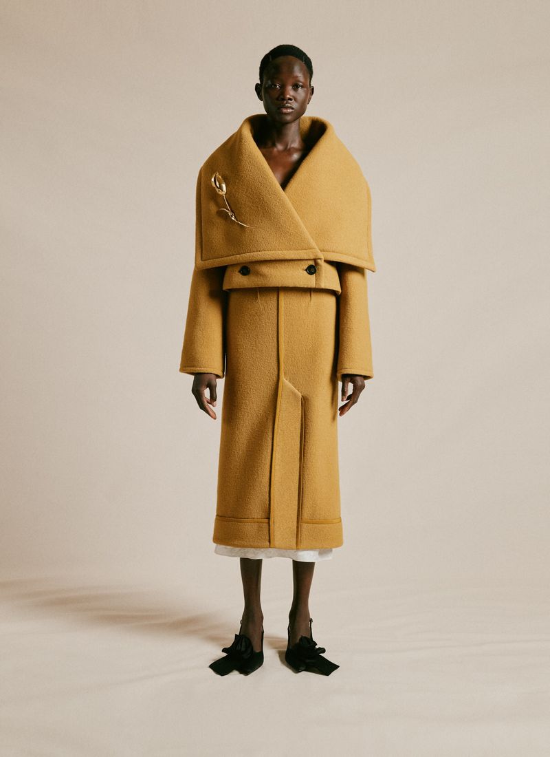 Fashionable Black woman poses in a draped, mustard-colored coat and flats in a studio setting, embodying elegance and contemporary style.