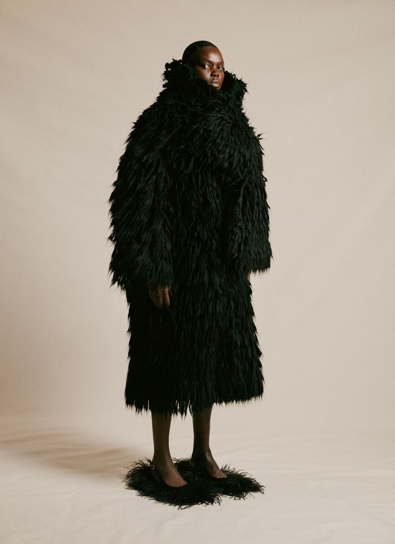 Fashionable woman wearing a large, textured black coat and matching shoes stands in a studio against a neutral background.