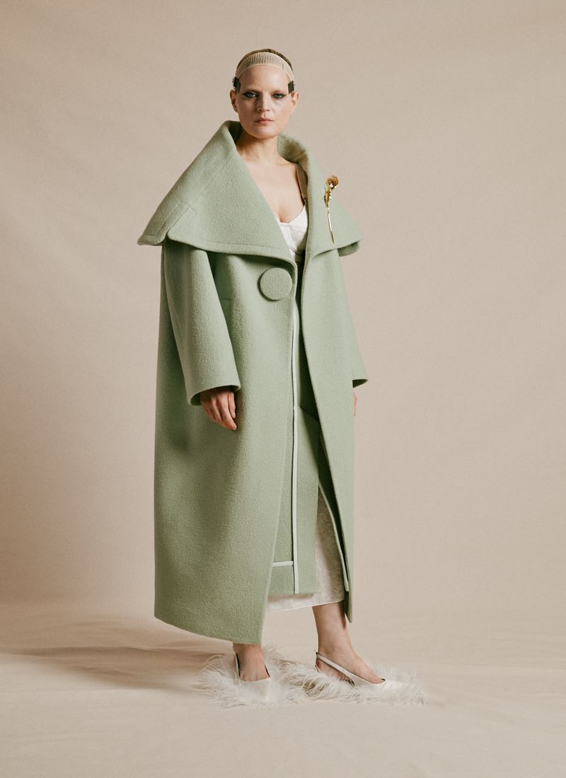 Woman with shaved head in a floor-length mint green coat with a large collar and skirt, posing indoors against a neutral background.