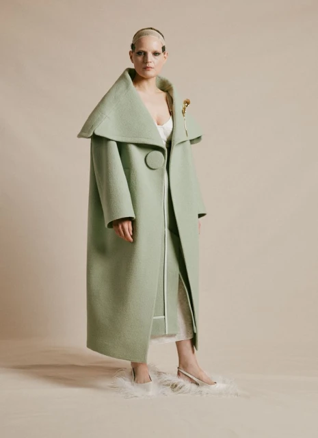 Woman with shaved head in a floor-length mint green coat with a large collar and skirt, posing indoors against a neutral background.