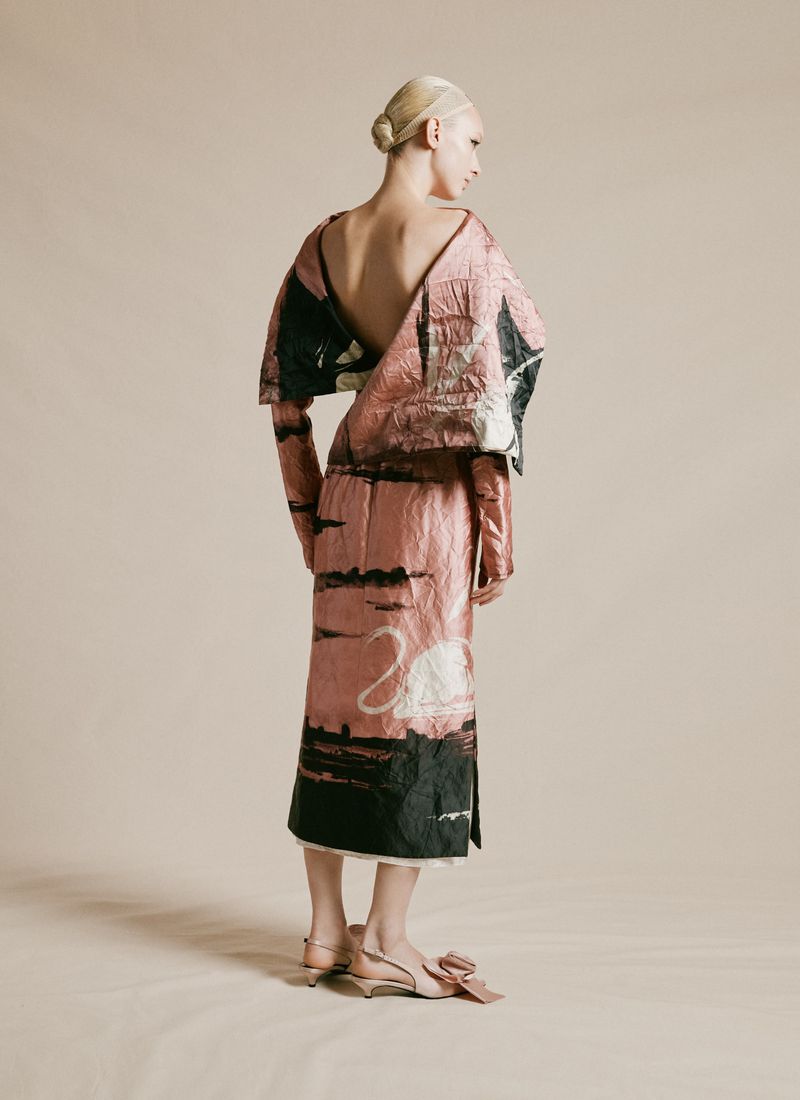 Back view of a slender adult female wearing a pink dress with swan print and slingback heels in a neutral-toned studio setting.