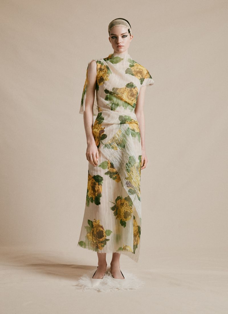 A slender woman poses in a translucent floral-print dress with a neutral backdrop, showcasing a modern and ethereal fashion aesthetic.