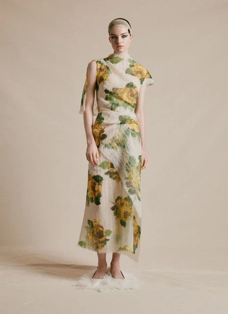 A slender woman poses in a translucent floral-print dress with a neutral backdrop, showcasing a modern and ethereal fashion aesthetic.