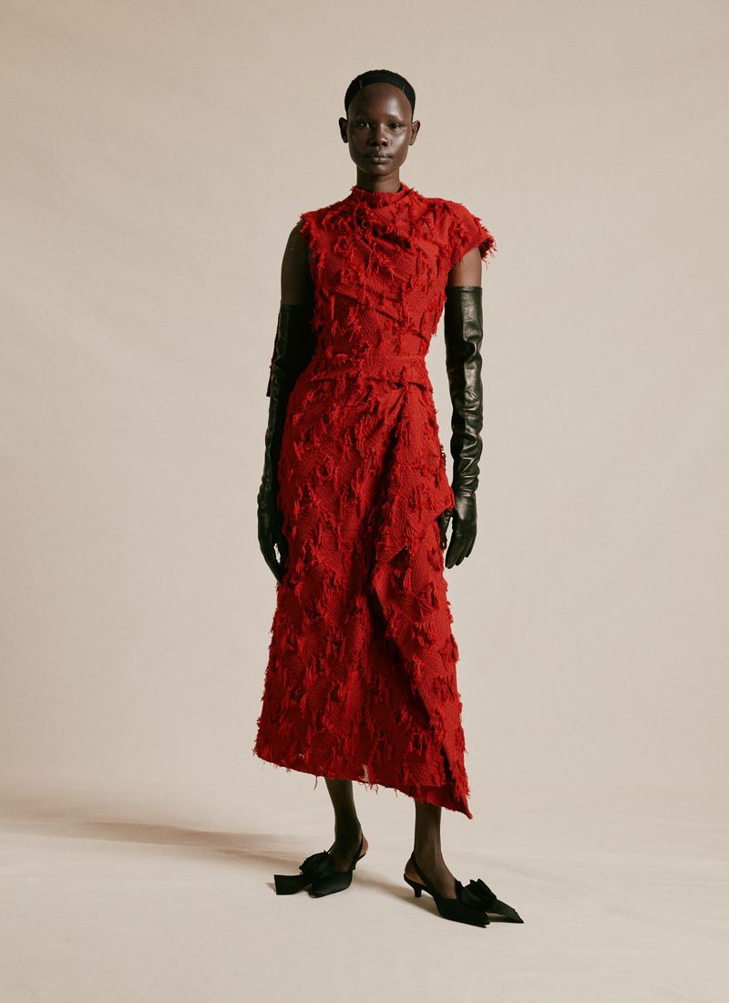 A tall, Black female model poses in a studio wearing a textured red dress with long black leather gloves and black kitten heels with bows.