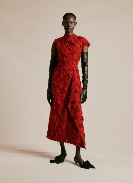 A tall, Black female model poses in a studio wearing a textured red dress with long black leather gloves and black kitten heels with bows.