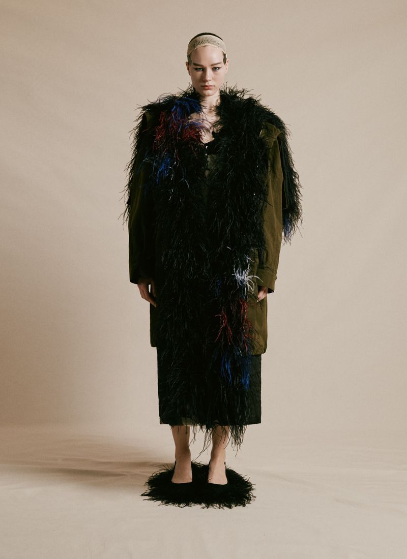 A model in feathered coat and heels poses in a studio with neutral backdrop, showcasing dramatic fashion and avant-garde style.