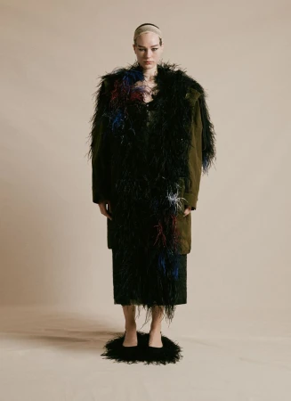 A model in feathered coat and heels poses in a studio with neutral backdrop, showcasing dramatic fashion and avant-garde style.