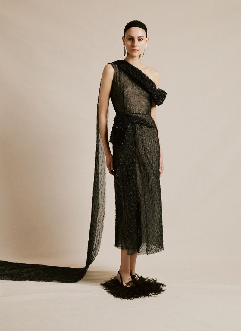A woman in an elegant black one-shoulder dress poses in a photo studio with a neutral background, creating a high-fashion editorial image.