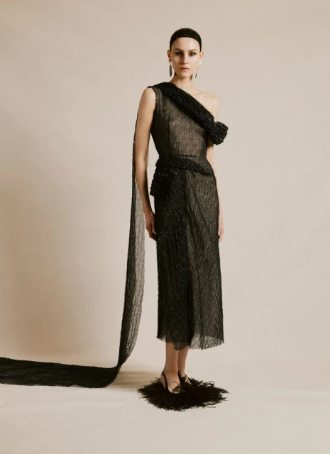 A woman in an elegant black one-shoulder dress poses in a photo studio with a neutral background, creating a high-fashion editorial image.