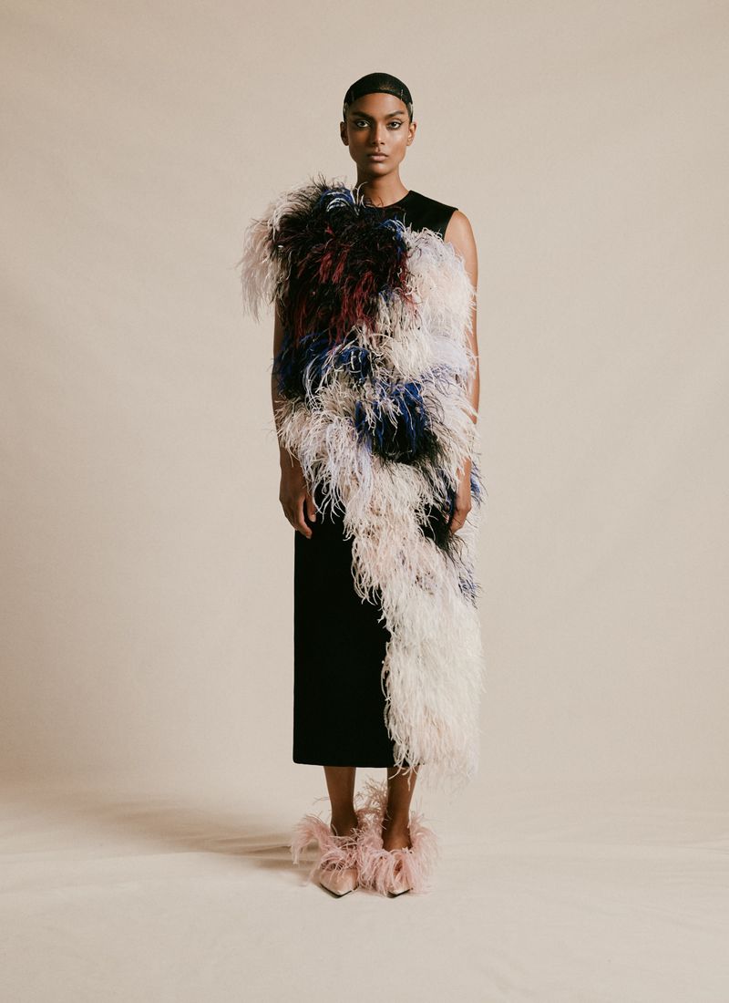 An adult female poses in a sleeveless black midi dress adorned with large, colorful feathers, and matching feather shoes, on a neutral backdrop.