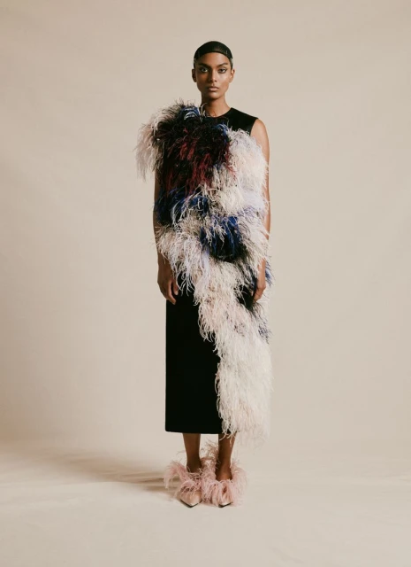 An adult female poses in a sleeveless black midi dress adorned with large, colorful feathers, and matching feather shoes, on a neutral backdrop.