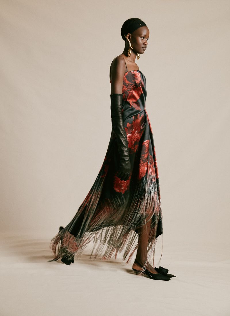Elegant Black woman in a red and black rose dress with a fringed hemline, long black gloves, and gold earrings on a plain background.