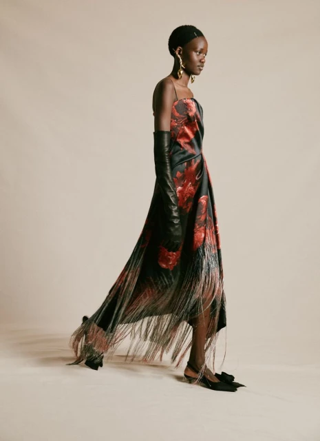 Elegant Black woman in a red and black rose dress with a fringed hemline, long black gloves, and gold earrings on a plain background.