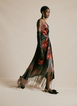 Elegant Black woman in a red and black rose dress with a fringed hemline, long black gloves, and gold earrings on a plain background.