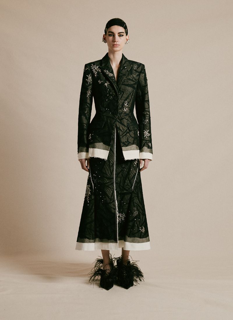 Full-length studio shot of a fashion model wearing a dark embellished jacket and skirt with feather shoes against a neutral background.
