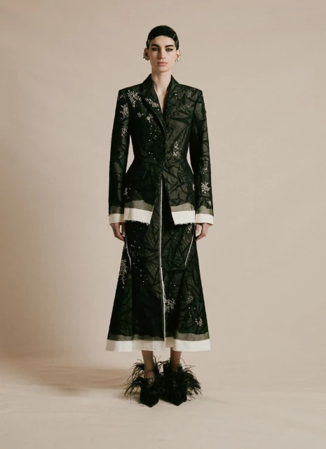 Full-length studio shot of a fashion model wearing a dark embellished jacket and skirt with feather shoes against a neutral background.