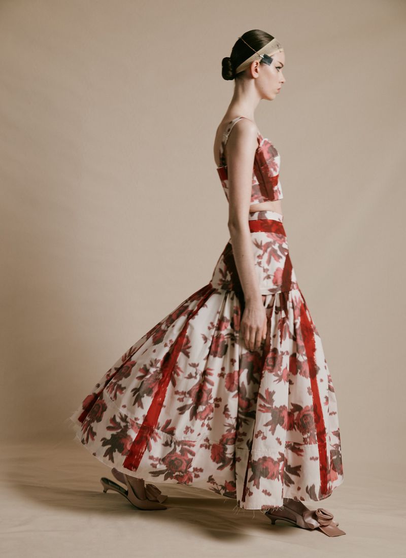 A woman in a floral two-piece dress with red accents poses in profile against a light background in a fashion photography studio setting.