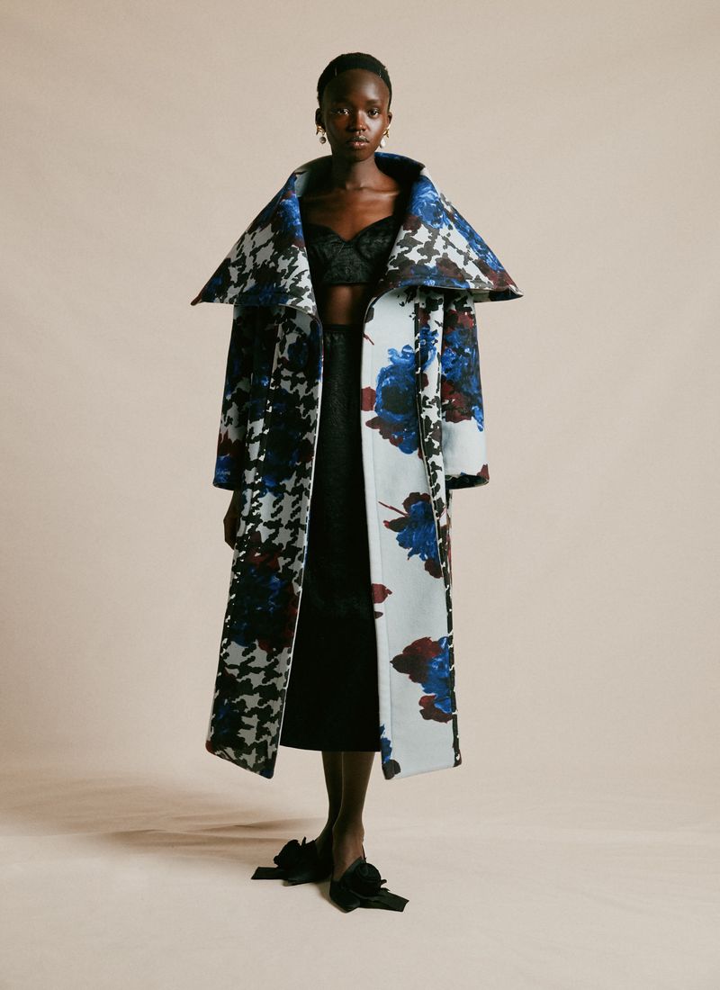 Fashion shot of a Black woman in a long, wide collared coat with floral and houndstooth patterns, a black skirt and bow-tied shoes.