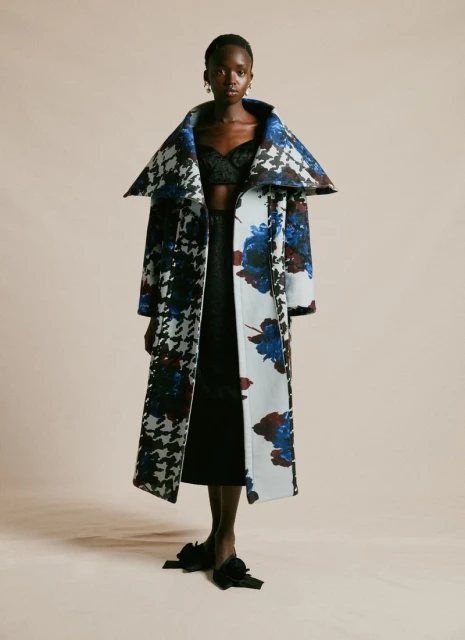 Fashion shot of a Black woman in a long, wide collared coat with floral and houndstooth patterns, a black skirt and bow-tied shoes.