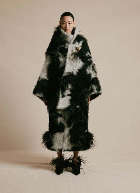 An Asian woman poses in a full-length shot, wearing an extravagant black and white patterned feather coat and matching shoes.