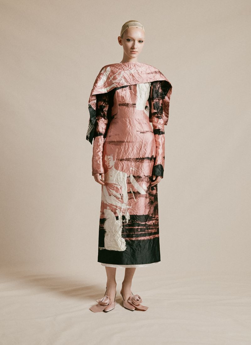 Fashion model wearing a pink, black, and white printed mid-length dress with a matching cape in a studio setting against a beige backdrop.