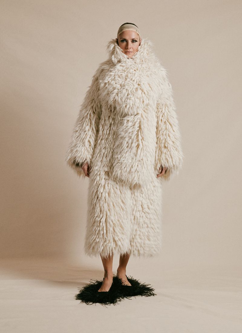 Full shot of a woman in a shaggy white coat and pants standing on a black, feathered mat in front of a beige backdrop.