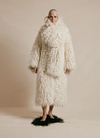Full shot of a woman in a shaggy white coat and pants standing on a black, feathered mat in front of a beige backdrop.