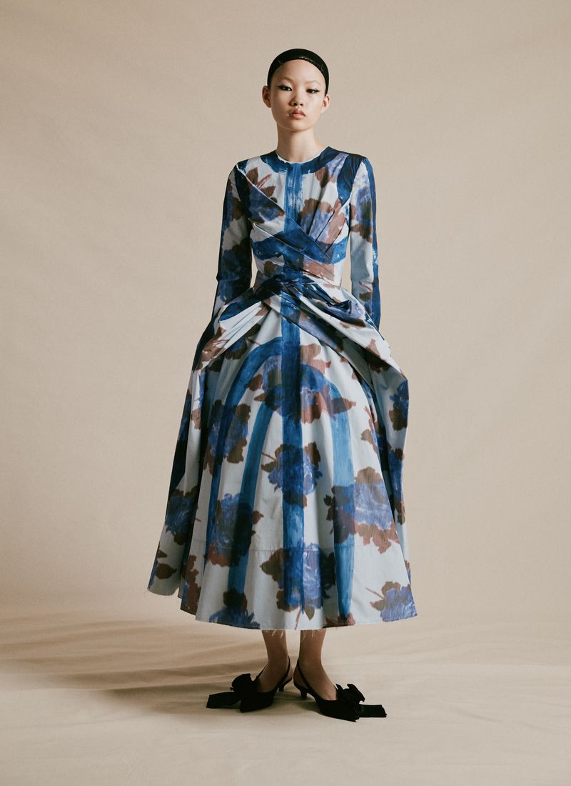 Asian woman in a full-length portrait wearing a blue and beige floral print dress and black heels in a neutral studio setting.