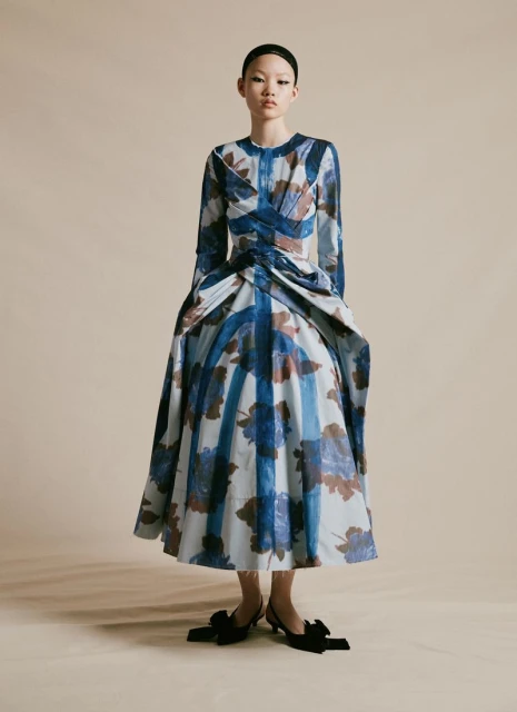 Asian woman in a full-length portrait wearing a blue and beige floral print dress and black heels in a neutral studio setting.