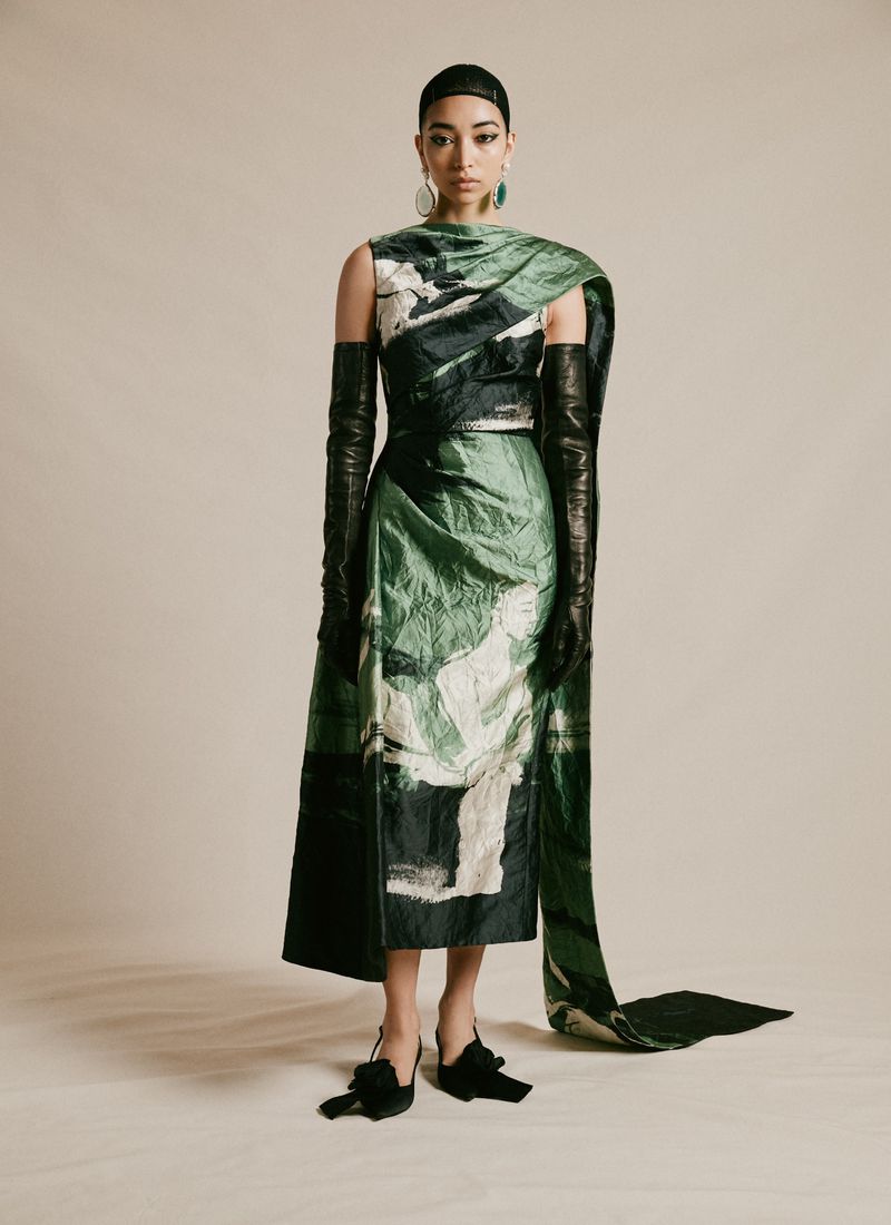An Asian woman poses in a green, white, and black sleeveless dress with long black leather gloves, black heels, and green teardrop earrings.