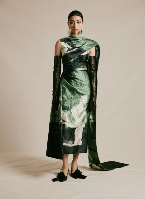 An Asian woman poses in a green, white, and black sleeveless dress with long black leather gloves, black heels, and green teardrop earrings.