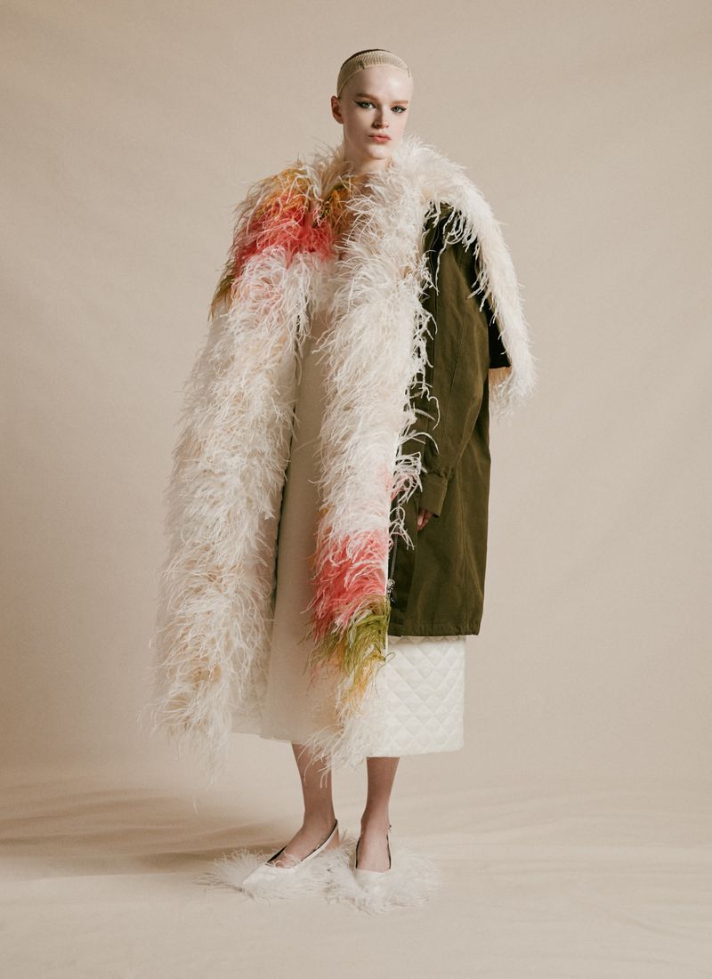 Full shot of a blonde woman in a white dress and feathered coat standing on a neutral background in a fashion-style portrait.