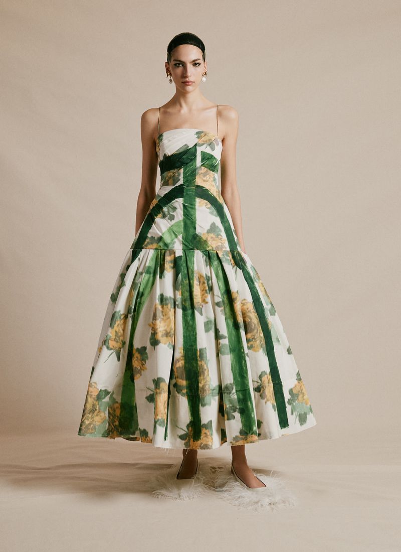 A woman in an elegant white floral dress stands indoors. The dress features yellow floral patterns and green vertical stripes.