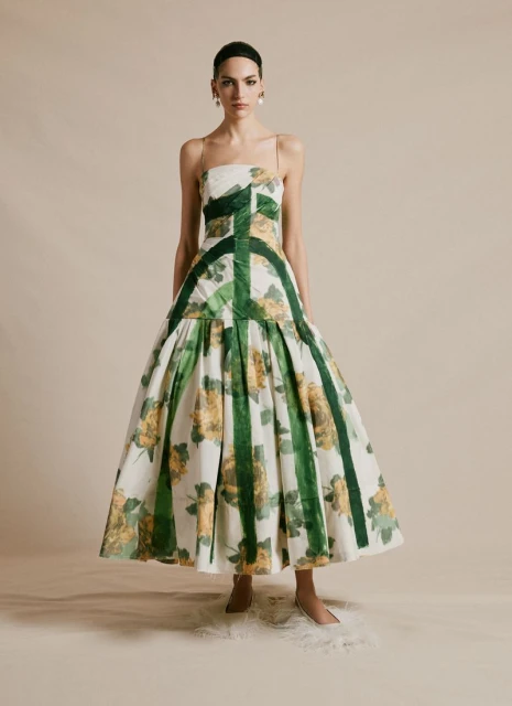 A woman in an elegant white floral dress stands indoors. The dress features yellow floral patterns and green vertical stripes.