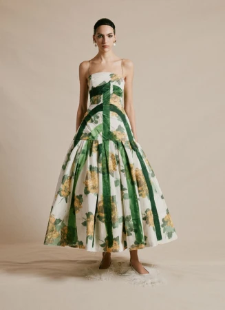 A woman in an elegant white floral dress stands indoors. The dress features yellow floral patterns and green vertical stripes.