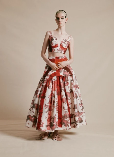 Fashion model poses in studio wearing floral crop top and skirt ensemble with brown accents on a neutral background in portrait orientation.