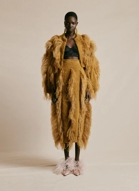 Full-length shot of a slender Black female model in a fuzzy, tan two-piece outfit with feathered shoes against a neutral backdrop.