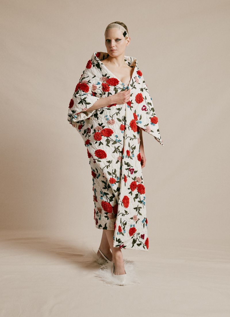 A model wearing a floral-patterned coat stands indoors against a neutral backdrop, showcasing high fashion and unique textures in a studio setting.
