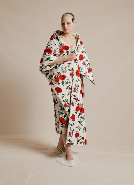 A model wearing a floral-patterned coat stands indoors against a neutral backdrop, showcasing high fashion and unique textures in a studio setting.