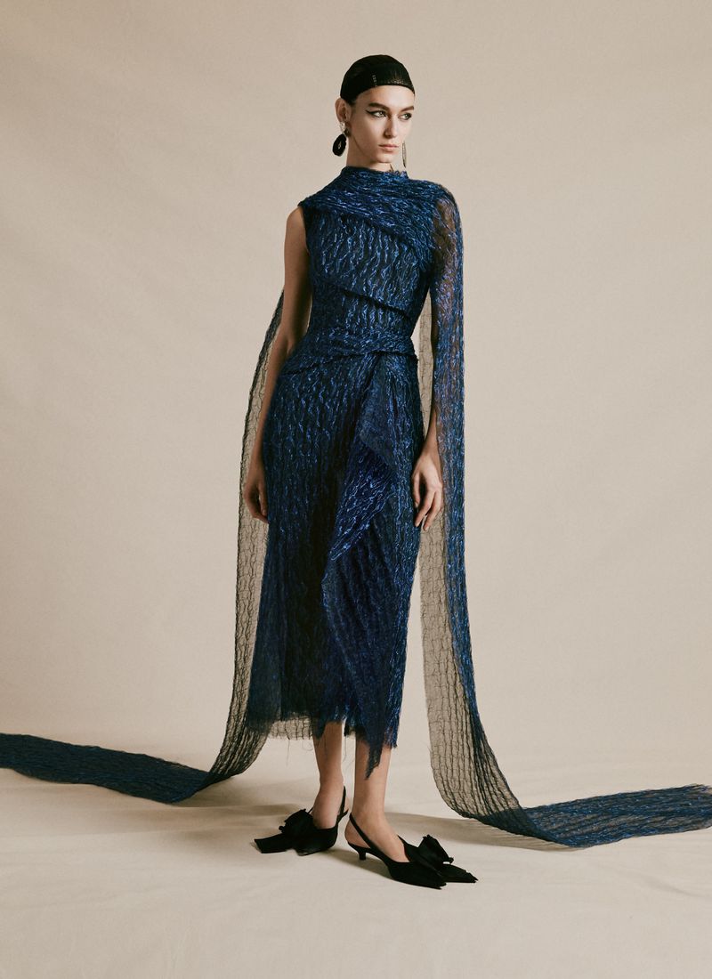 Woman poses in a textured blue midi dress with a draped shawl, complemented by black kitten heels with bows, against a light backdrop.