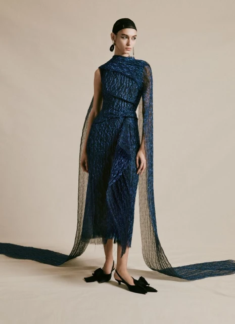 Woman poses in a textured blue midi dress with a draped shawl, complemented by black kitten heels with bows, against a light backdrop.