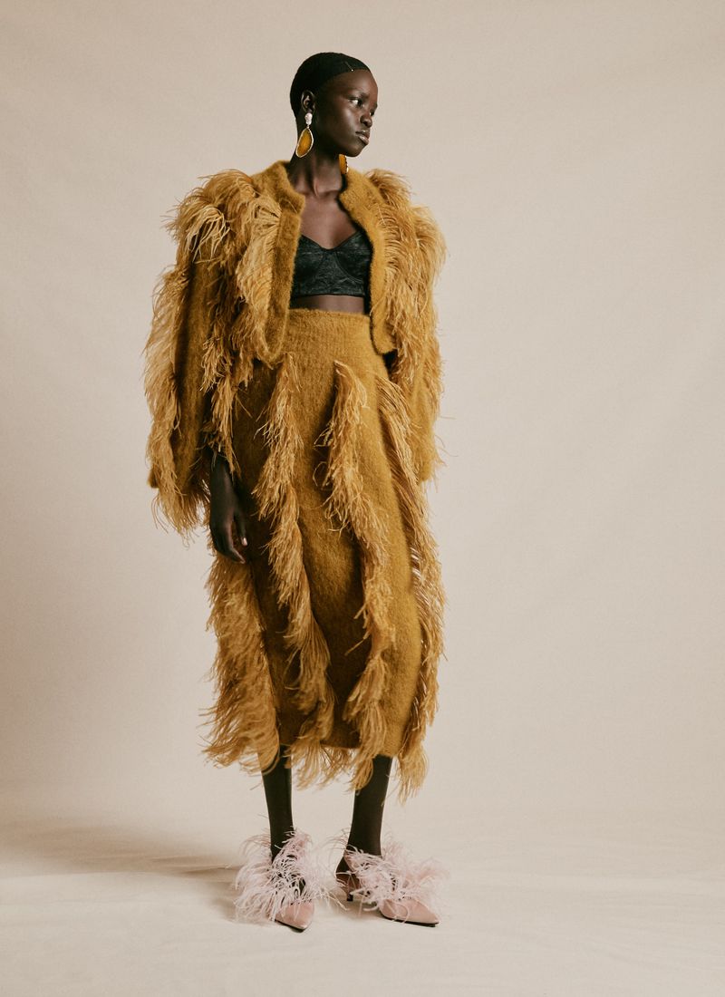 Full-length portrait of a Black female fashion model in a feathered jacket and skirt, accessorized with large earrings and feathered heels.