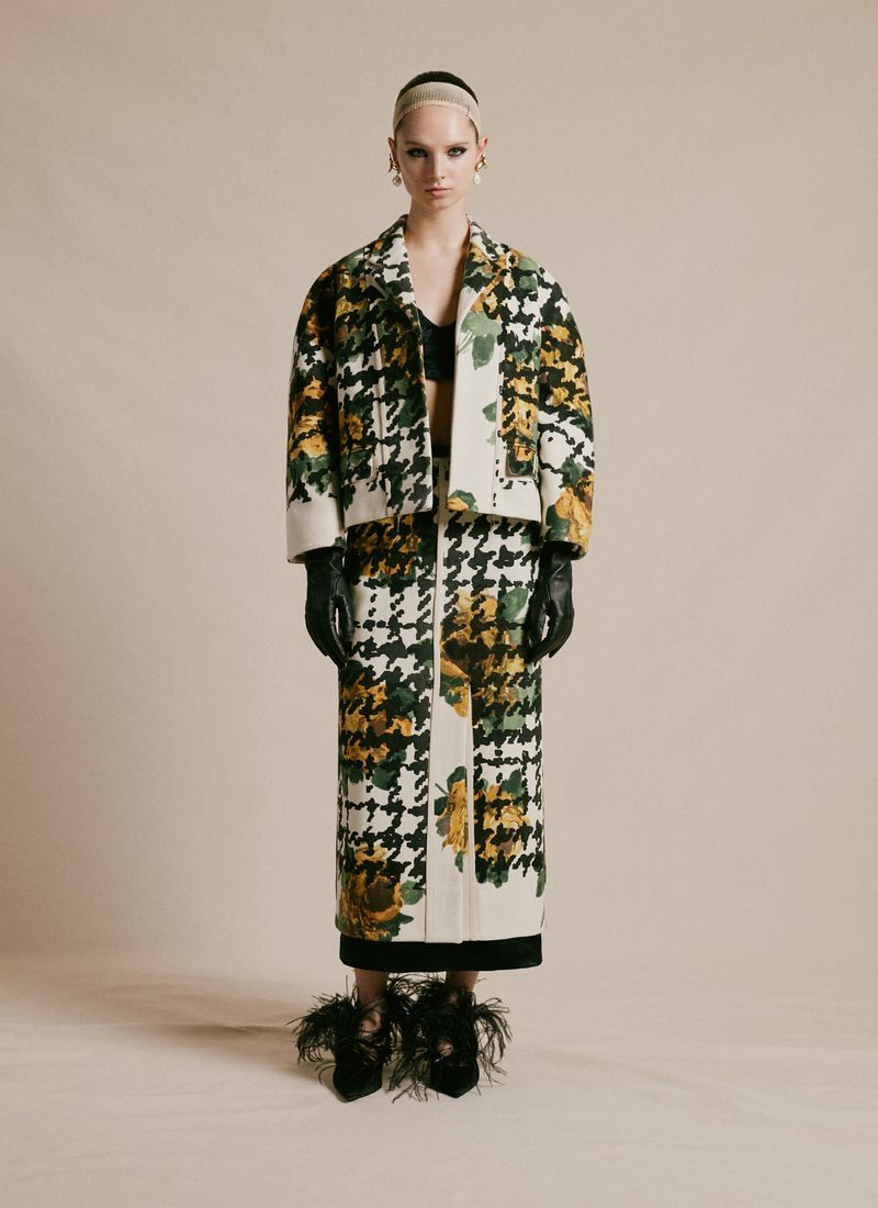 Full-shot of a female model wearing a houndstooth and floral print jacket and skirt, paired with feather-embellished heels in a studio.