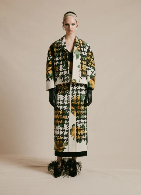 Full-shot of a female model wearing a houndstooth and floral print jacket and skirt, paired with feather-embellished heels in a studio.