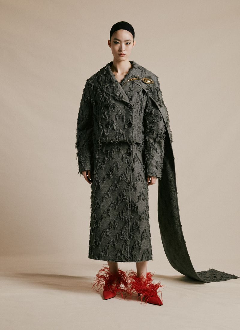 An Asian female model is posing in a photo studio, wearing a frayed gray jacket, a skirt, and red feathered high heels in a full shot.