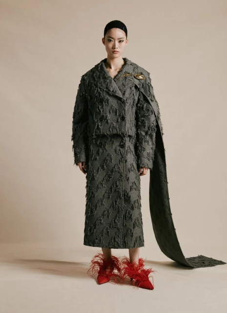 An Asian female model is posing in a photo studio, wearing a frayed gray jacket, a skirt, and red feathered high heels in a full shot.