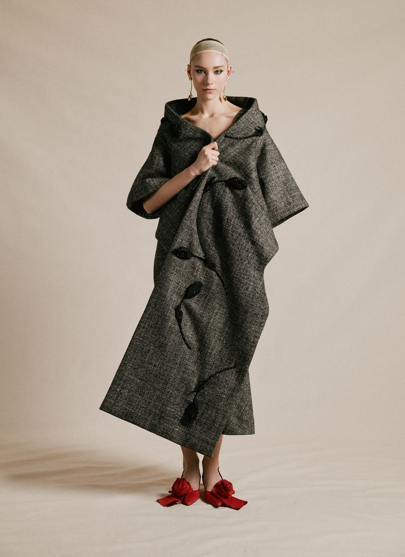 Fashion model posing in oversized gray hooded coat and skirt with red floral shoes in a neutral studio setting under studio lighting.